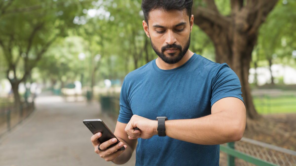 Runner in a park checking heart-rate graphs on a smartwatch, smart ring on finger, phone in hand