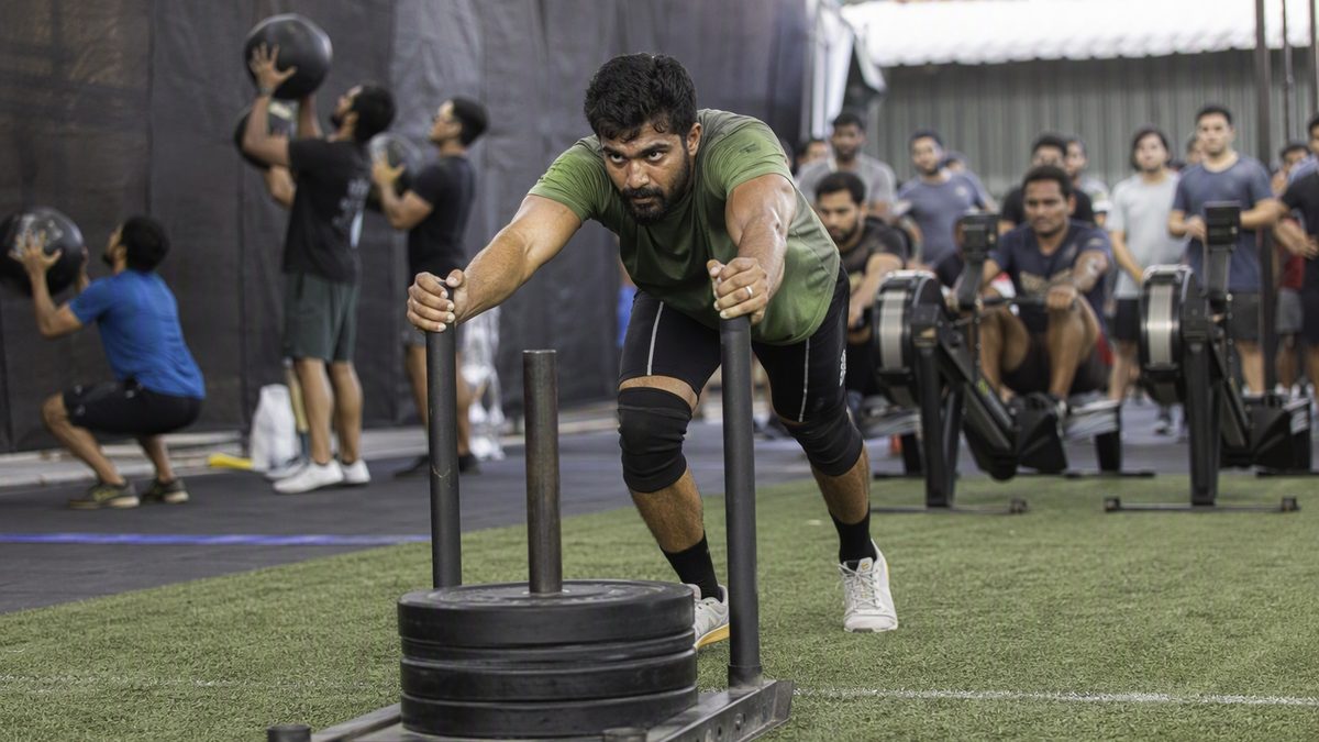 Athlete pushing a weighted sled on turf in an indoor fitness race venue, wall balls and rowers in the background